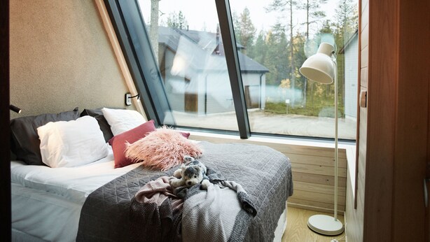 Une chambre avec une grande fenêtre, un lit avec des oreillers et une couverture, et une lampe de chevet.