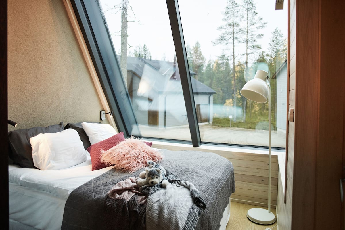 Een slaapkamer met een groot raam, een bed met kussens en een deken, en een bedlamp.
