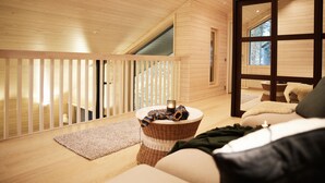Une pièce chaleureuse aux murs et au plafond en bois, un balcon avec une balustrade, une table en osier avec des bougies et un canapé recouvert d’une couverture.