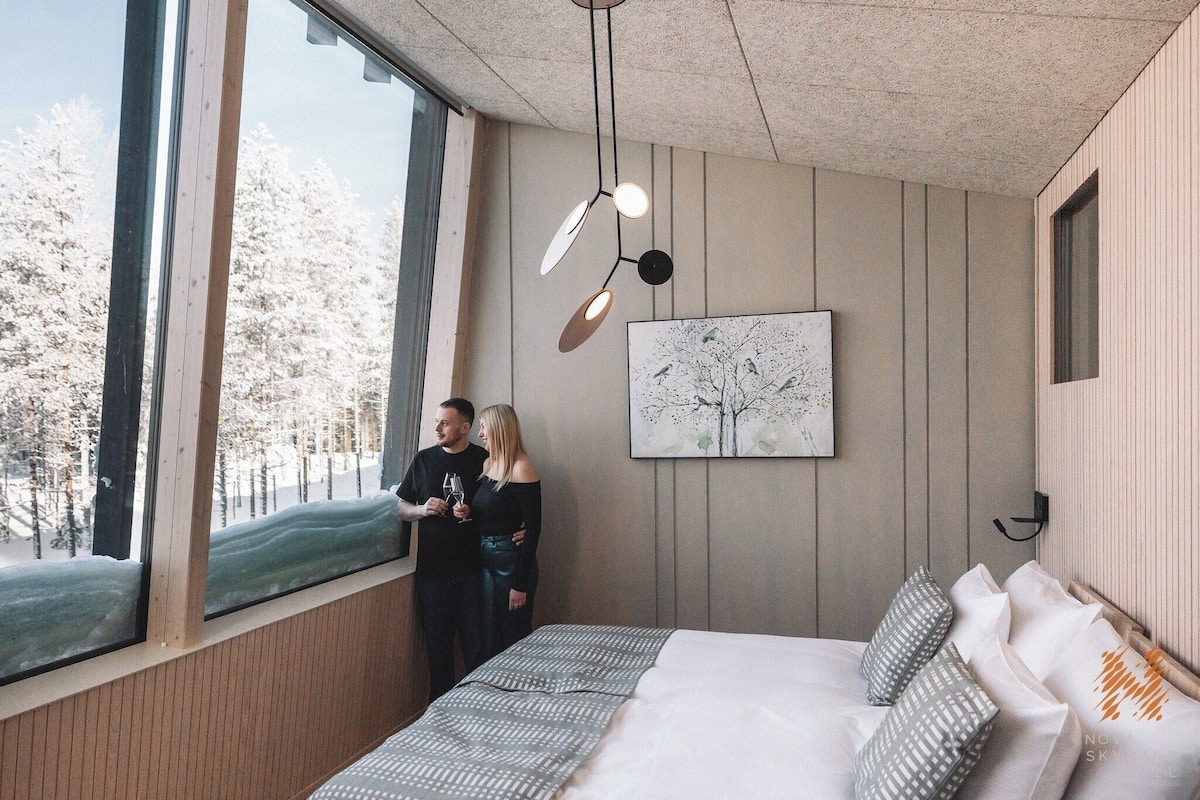 Een slaapkamer met een groot raam, een bed met beddengoed met een patroon, een hanglamp en een aan de muur hangend schilderij.