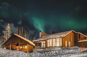 Una cabaña de madera con ventanas grandes, rodeada de nieve, bajo un cielo nocturno con auroras boreales visibles.