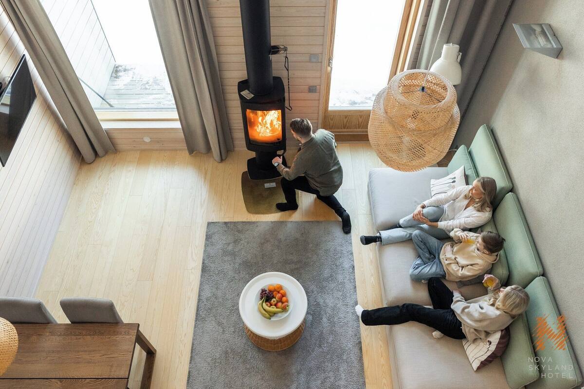 Een moderne woonkamer met een houtkachel, een zithoek, een eettafel met fruit en een groot raam.
