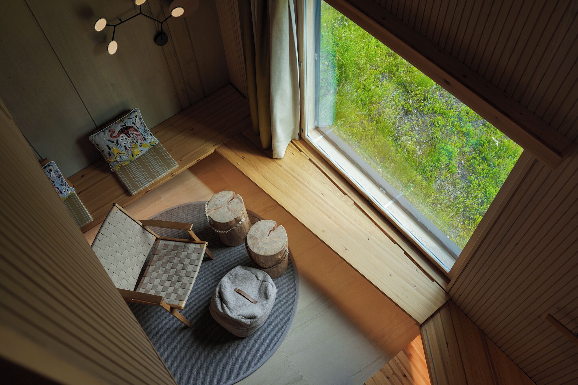 Een gezellige kamer met een houten vloer, een ronde zithoek met een geweven mat, een kleine tafel met een decoratief kussen en uitzicht op groen buiten.