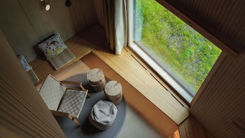Een gezellige kamer met een houten vloer, een ronde zithoek met een geweven mat, een kleine tafel met een decoratief kussen en uitzicht op groen buiten.
