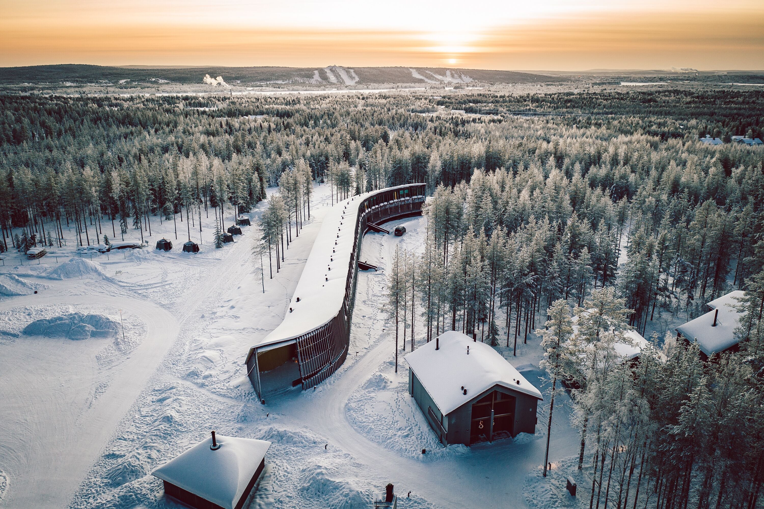 A snow-covered landscape with a unique curved building structure, surrounded by dense forest and a clear sky.