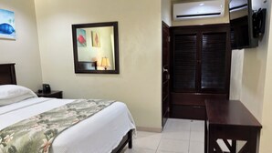 A hotel room with a bed, a desk, a chair, a wardrobe, and a wall-mounted TV. - Rainbow Hotel (Caye Caulker)