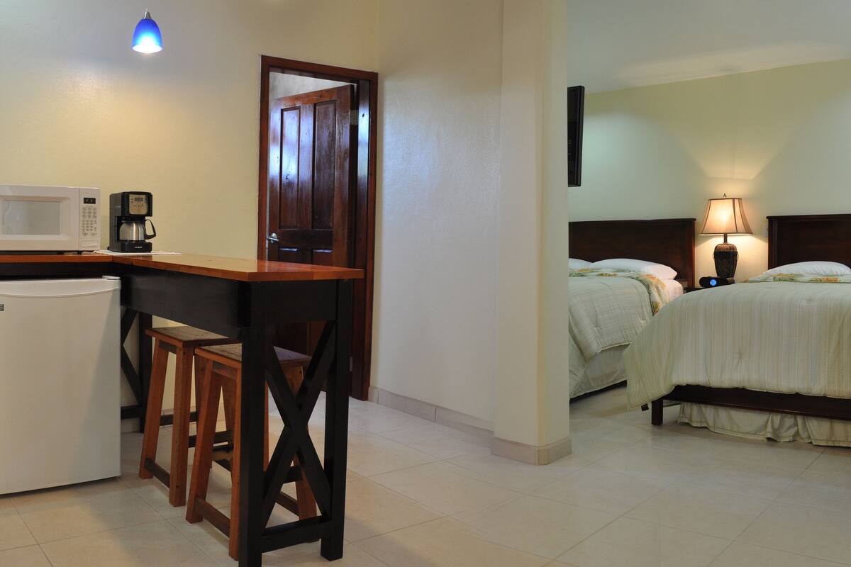 A compact hotel room with a kitchenette, a bed with two pillows, and a bedside lamp.