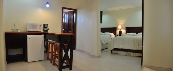 A compact hotel room with a kitchenette, a bed with two pillows, and a bedside lamp. - Rainbow Hotel (Caye Caulker)