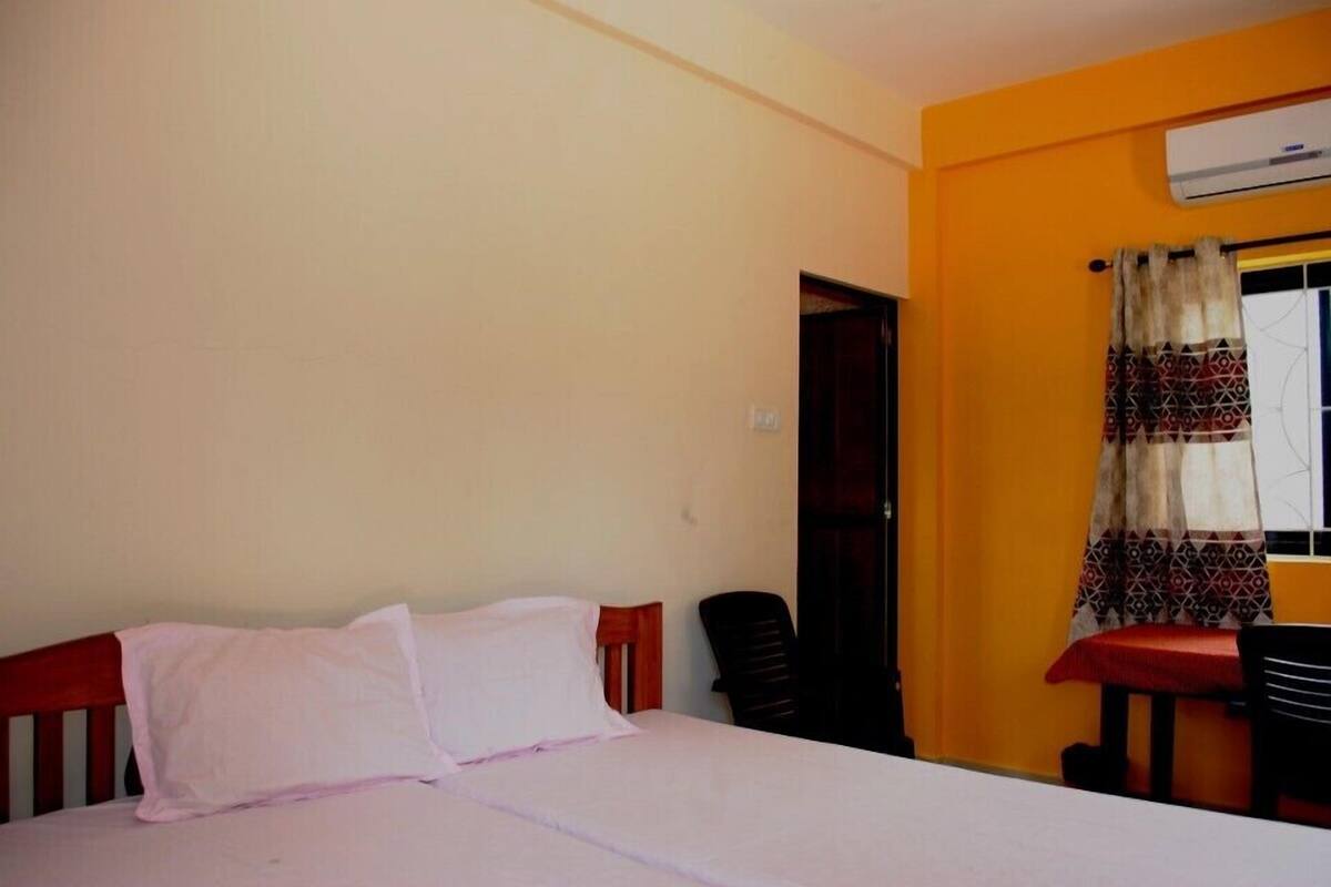A single bed with a wooden headboard, a small table with a red cushion, and a chair.