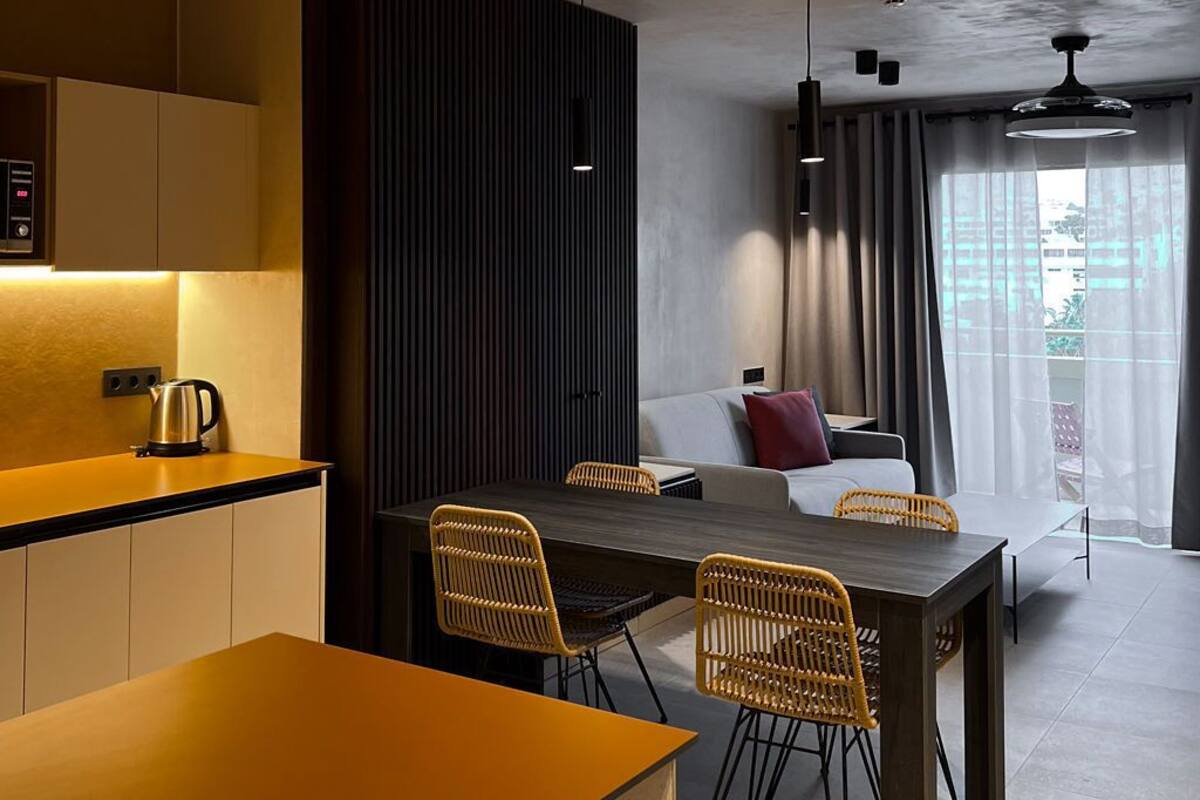 A modern kitchen with a wooden countertop, a dining area with a dark table and wicker chairs, and a living area with a sofa and curtains.