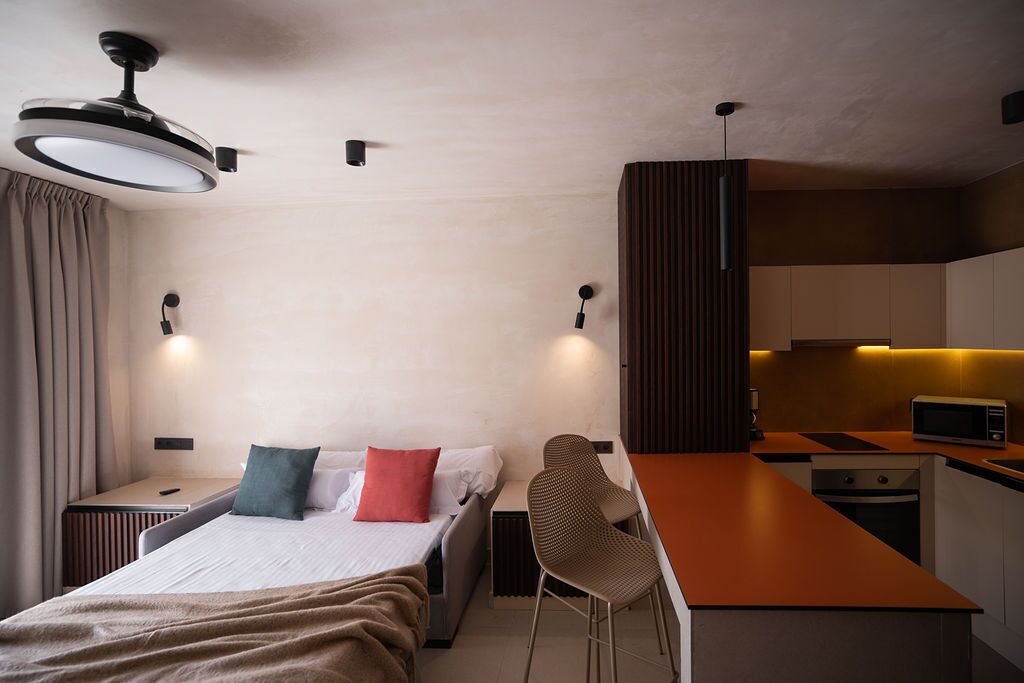 Een moderne hotelkamer met een bed, nachtkastjes, een bureau, een stoel, een kitchenette en een plafondventilator.