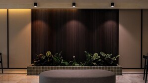 A modern waiting area with a round grey sofa, a tiled planter with green plants, and a dark wooden panel wall with vertical lines. - Apartamentos Playazul (Arona)