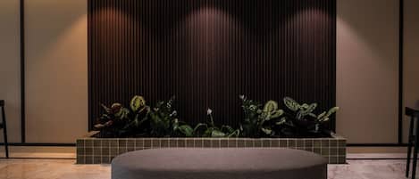 A modern waiting area with a round grey sofa, a tiled planter with green plants, and a dark wooden panel wall with vertical lines.