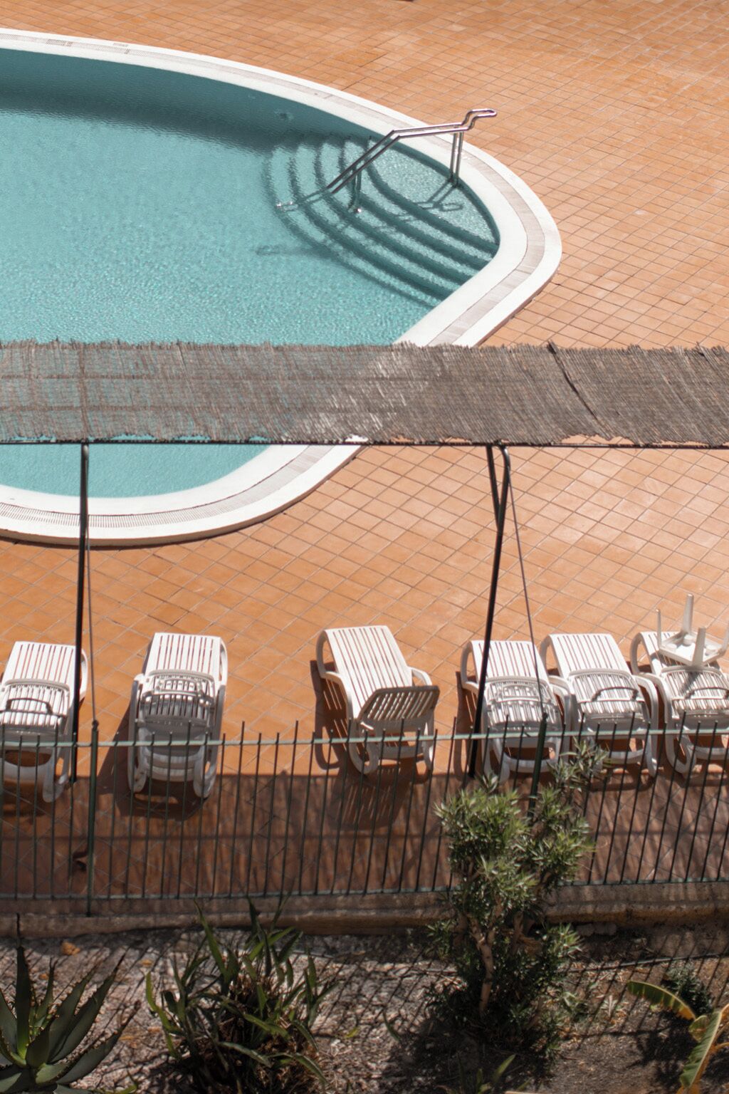 Una piscina con borde de baldosas, rodeada de sillas de descanso y una valla.