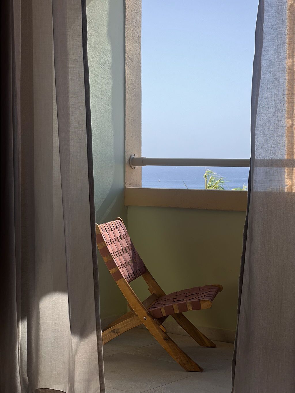 Un balcón con vista al mar, una silla de madera y cortinas translúcidas.