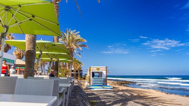 A beachside restaurant with outdoor seating, palm trees, and a view of the ocean. - Apartamentos Playazul (Arona)