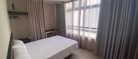 Habitación de hotel con cama individual, escritorio y silla. Tiene una ventana con cortinas y un perchero con ganchos.