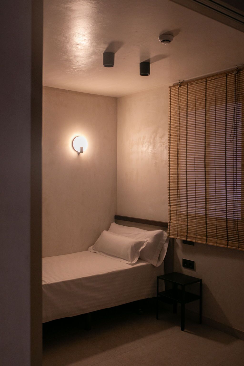 Une petite pièce faiblement éclairée, avec un lit simple, une lampe fixée au mur et une fenêtre munie de stores.