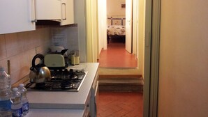 A kitchen with a gas stove, kettle, and water bottles. A hallway leads to a bedroom with a bed and a nightstand. - Appartamento Dante (Florence)