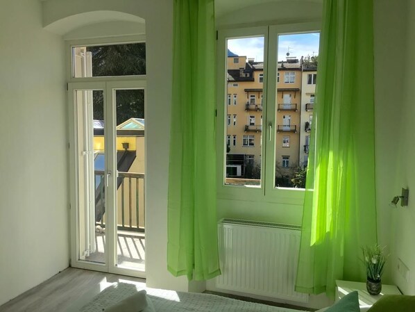 Habitación con balcón, ventana con cortinas verdes y vista a edificios.