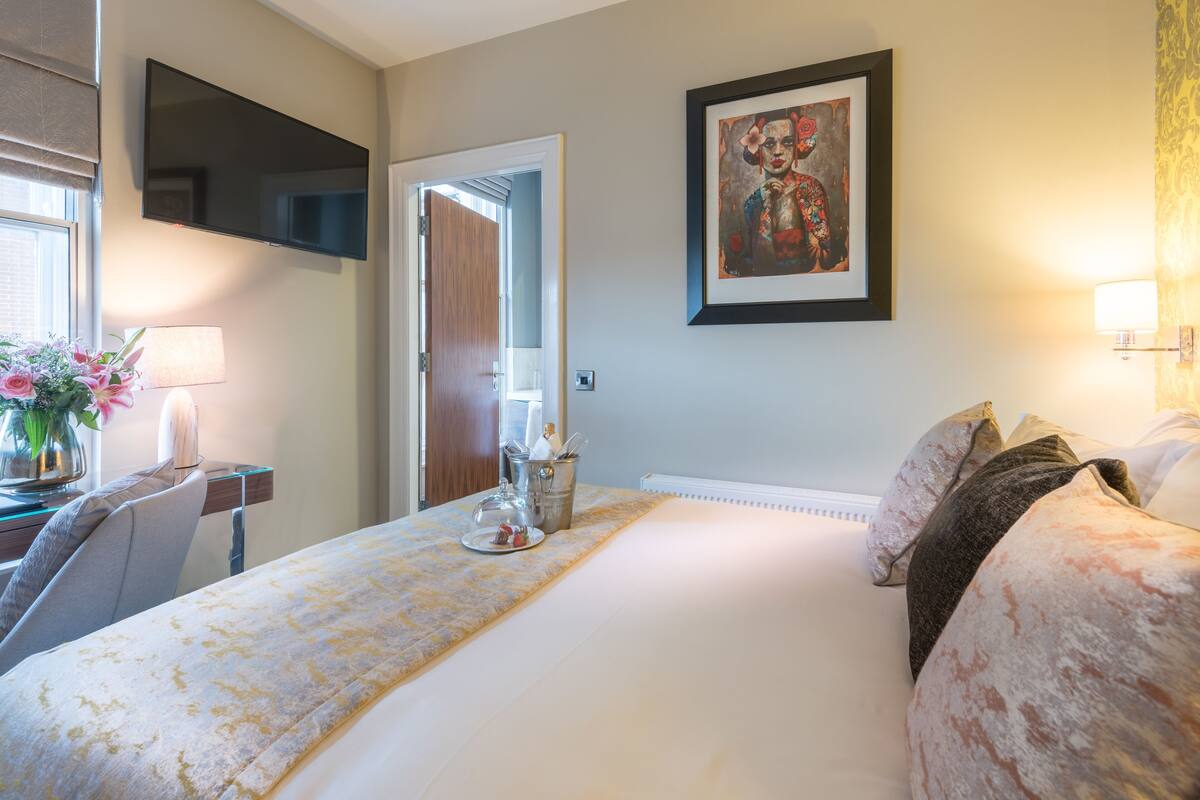 Habitación de hotel con cama, escritorio con lámpara, televisor y un cuadro en la pared.
