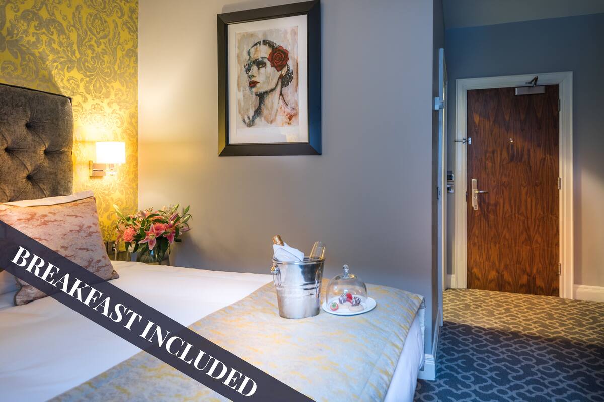 Habitación de hotel con cama, una obra de arte enmarcada en la pared, mesita de noche con lámpara, un jarrón con flores y una bandeja de desayuno sobre la cama.