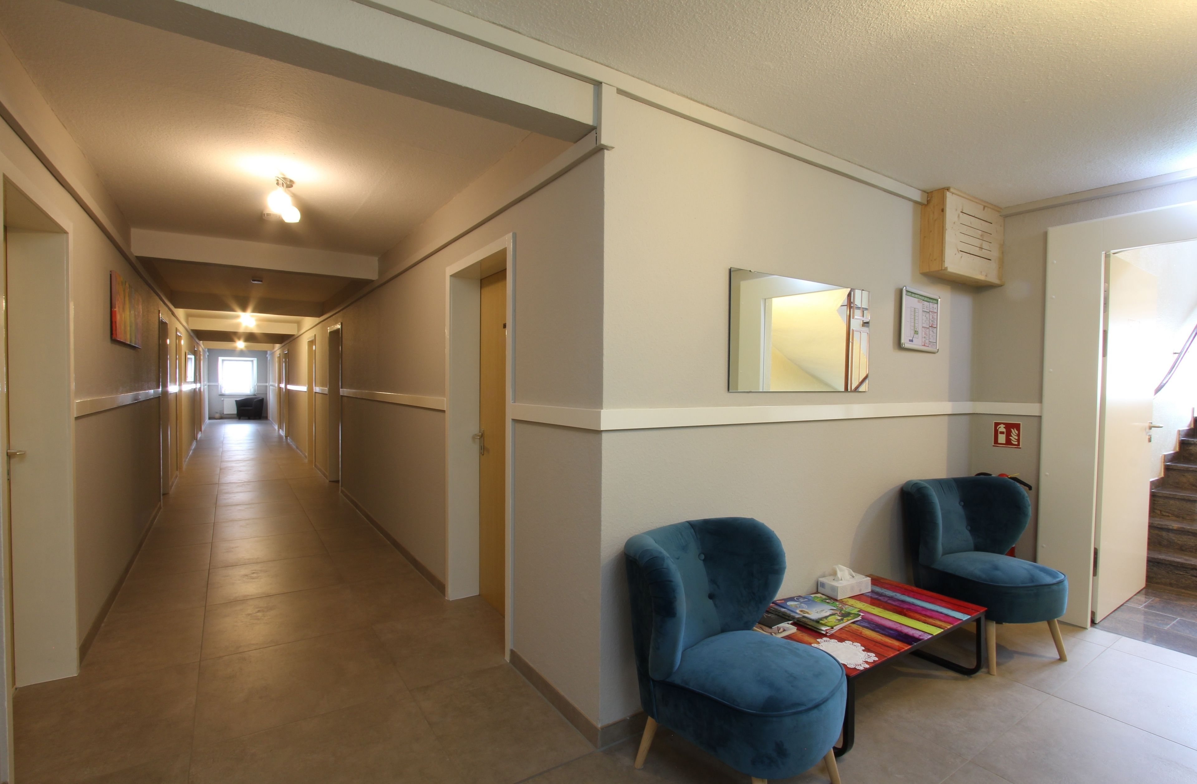 Een hotelgang met twee blauwe fauteuils, een kleurrijke streep tafel en een spiegel aan de muur.