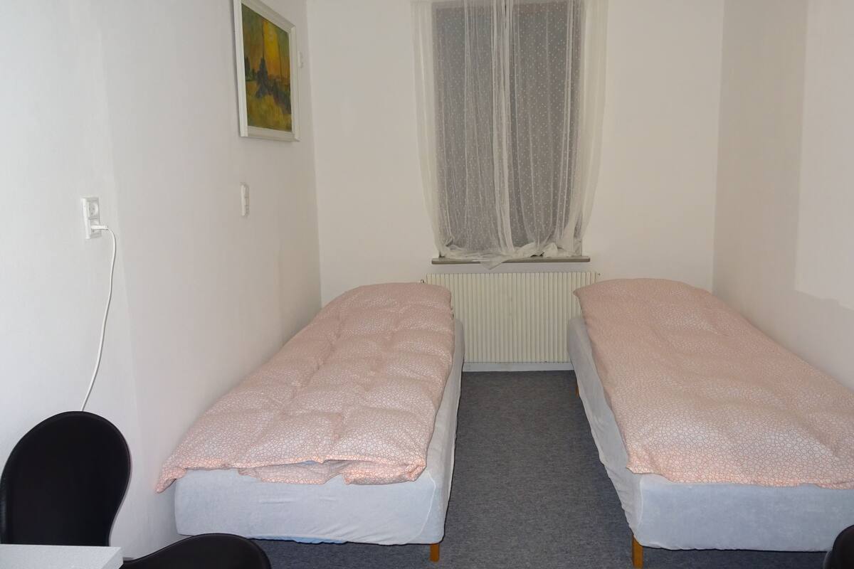 A small, clean room with two single beds, a window with sheer curtains, a radiator, and a painting on the wall.