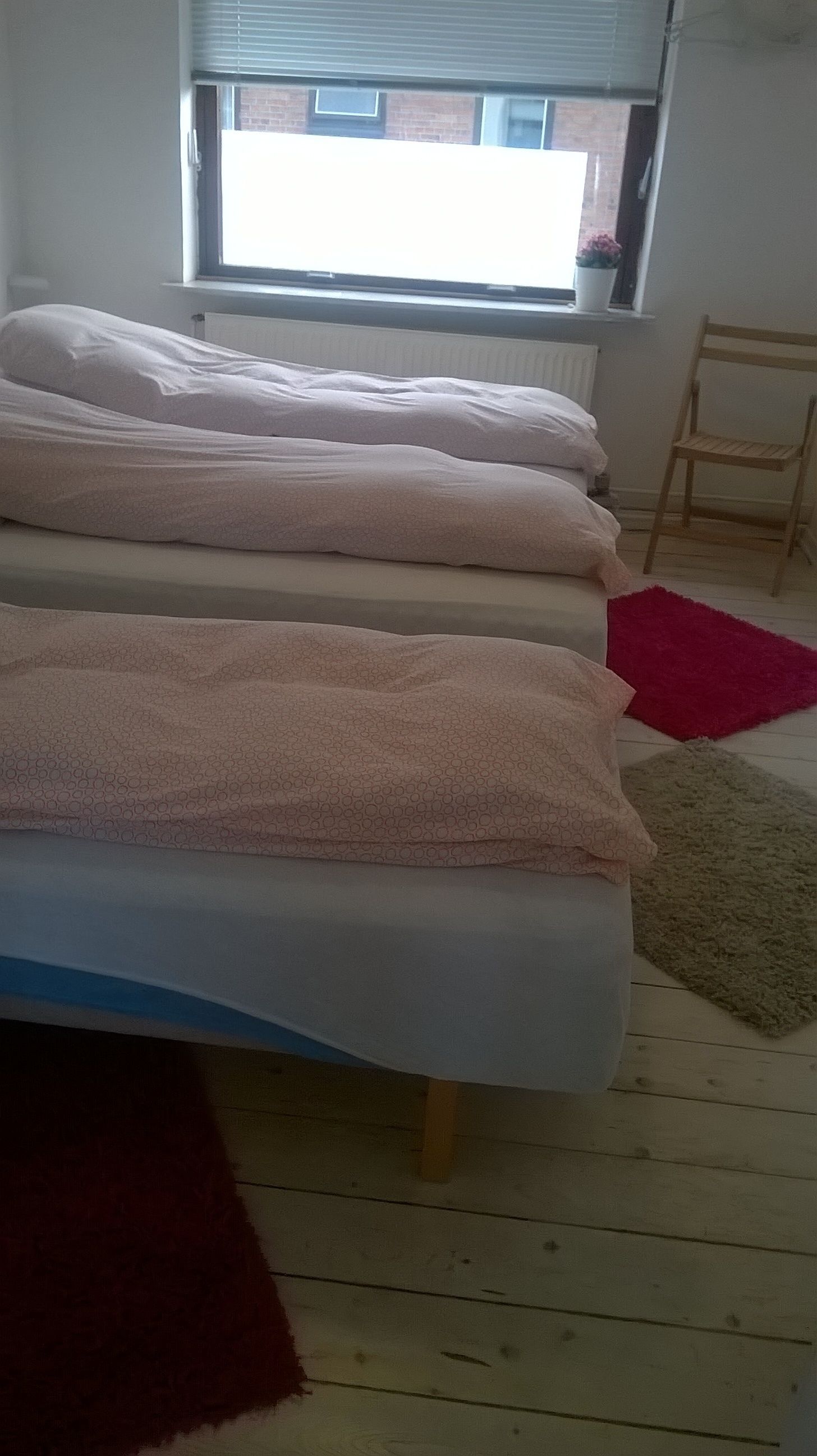 Un lit recouvert d’une couverture à motifs, une fenêtre avec des stores et une chaise dans une pièce dont le sol est en bois.