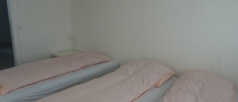 A room with two single beds, each with a pink blanket and a grey mattress. There is a white wall and a door visible.