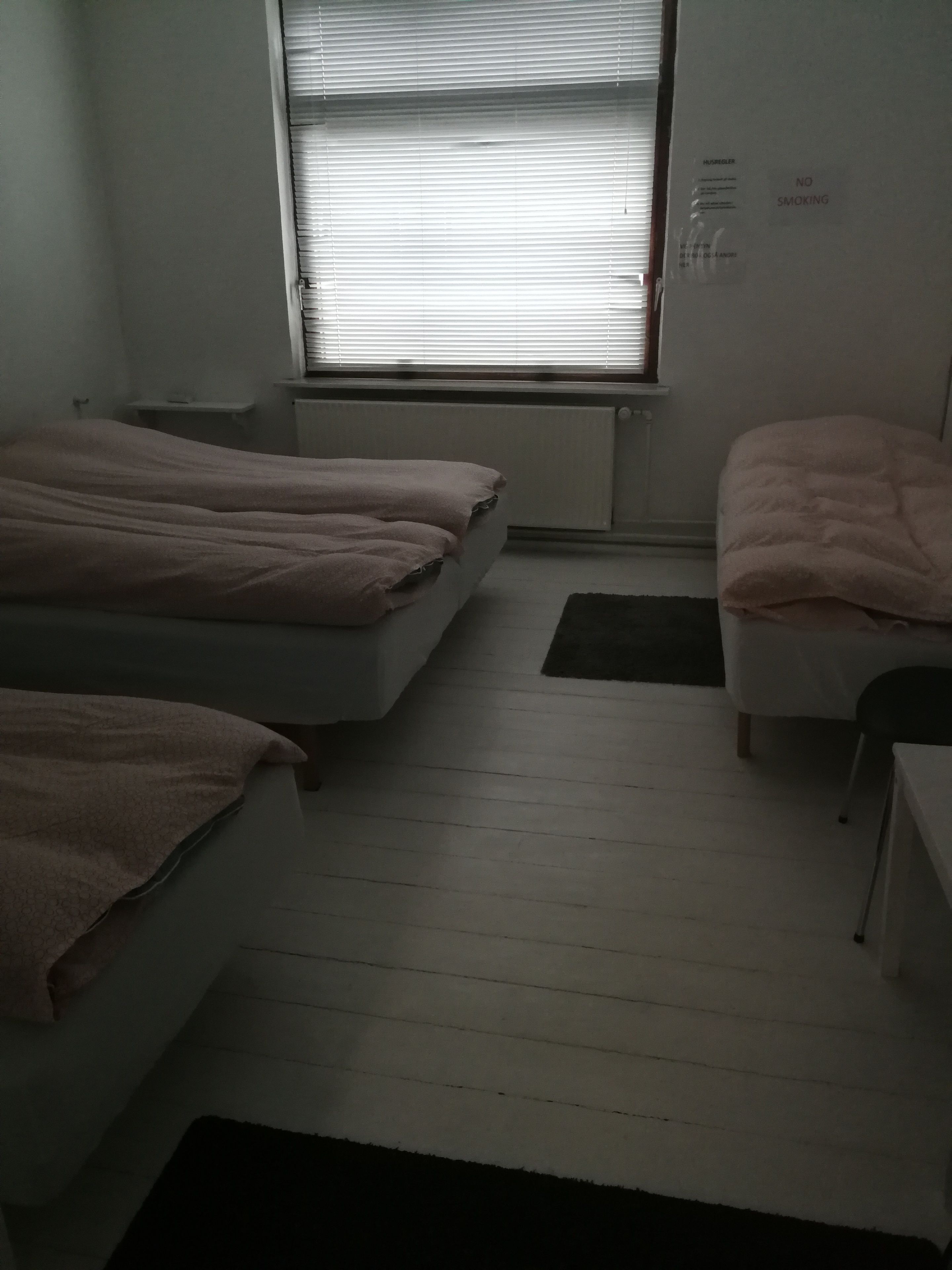A small, dimly lit room with two beds, a desk, and a radiator.