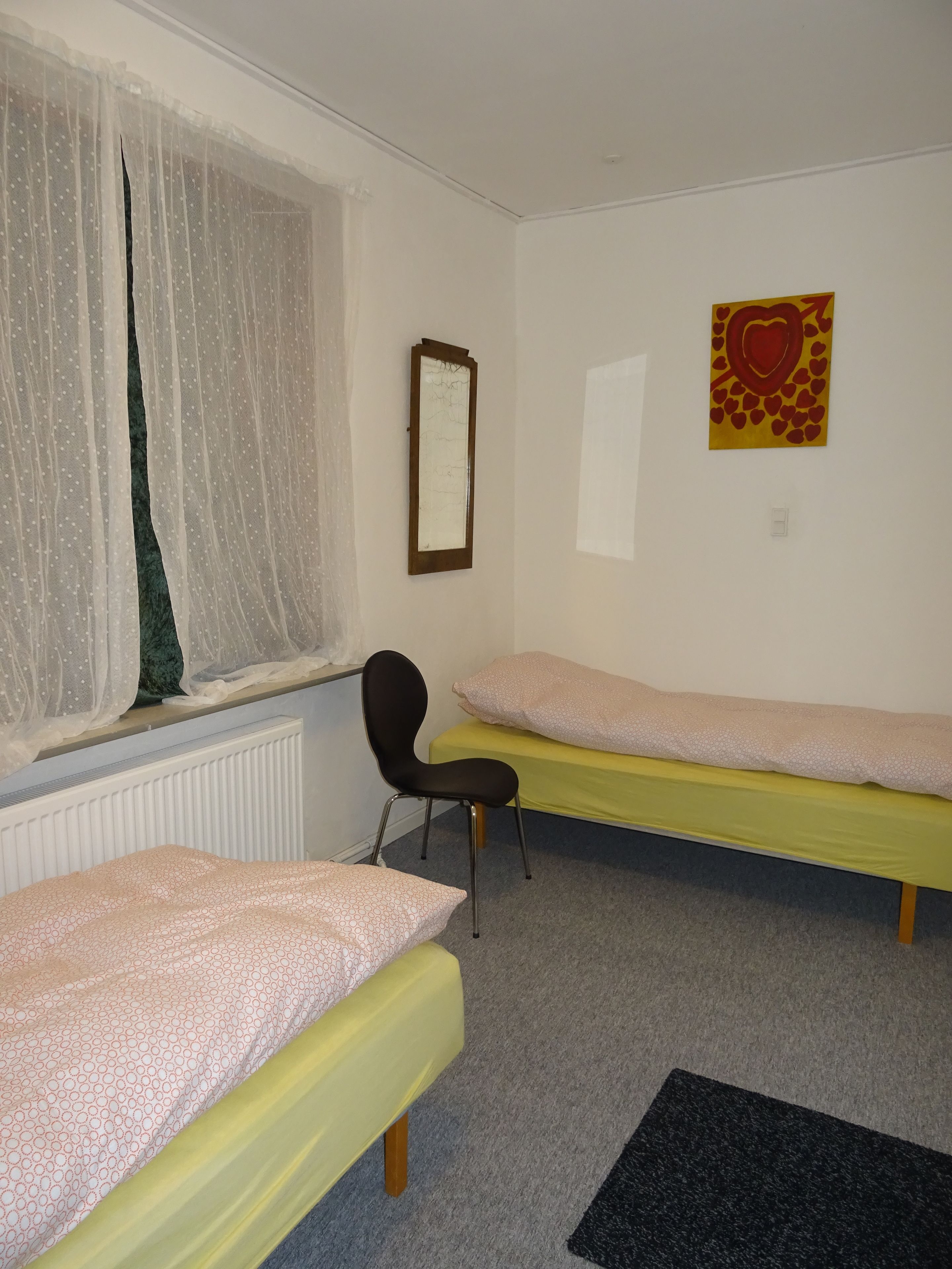 A room with two single beds, a chair, a radiator, a window with sheer curtains, and a wall with a heart-shaped artwork.