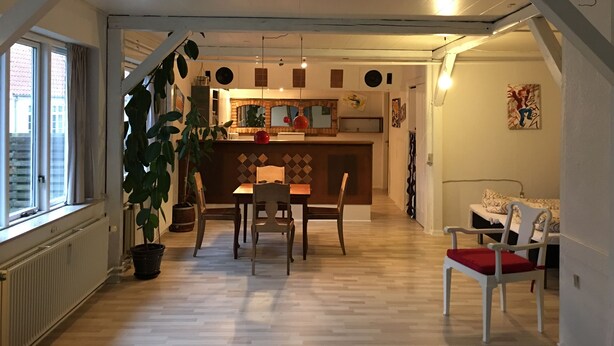 En matplats med ett träbord och stolar, en krukväxt och ett kök i bakgrunden.