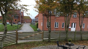 En innergård med en kullerstensgång som leder fram till en träport, omgiven av byggnader i tegel och träd.