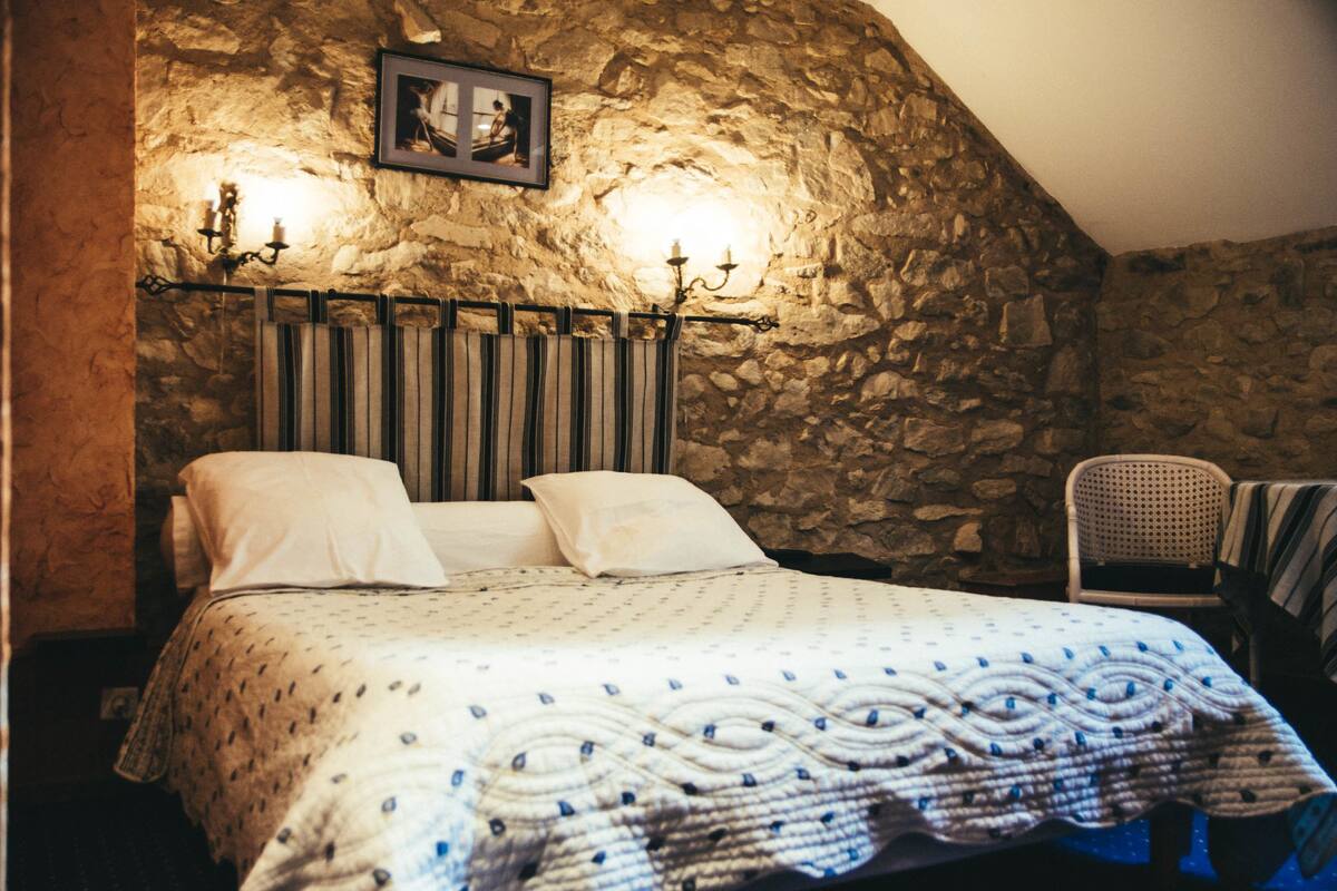 Une chambre avec un mur en pierre, un lit recouvert d’une couvre-lit à motifs, une chaise et un tableau accroché au mur.