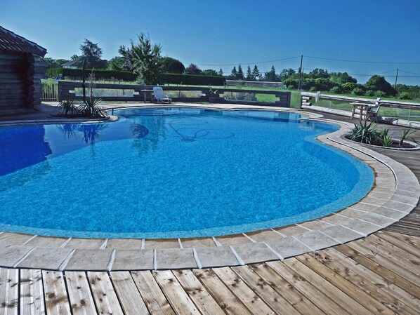 A swimming pool with a wooden deck surrounding it, a stone wall, and a view of the surrounding landscape.