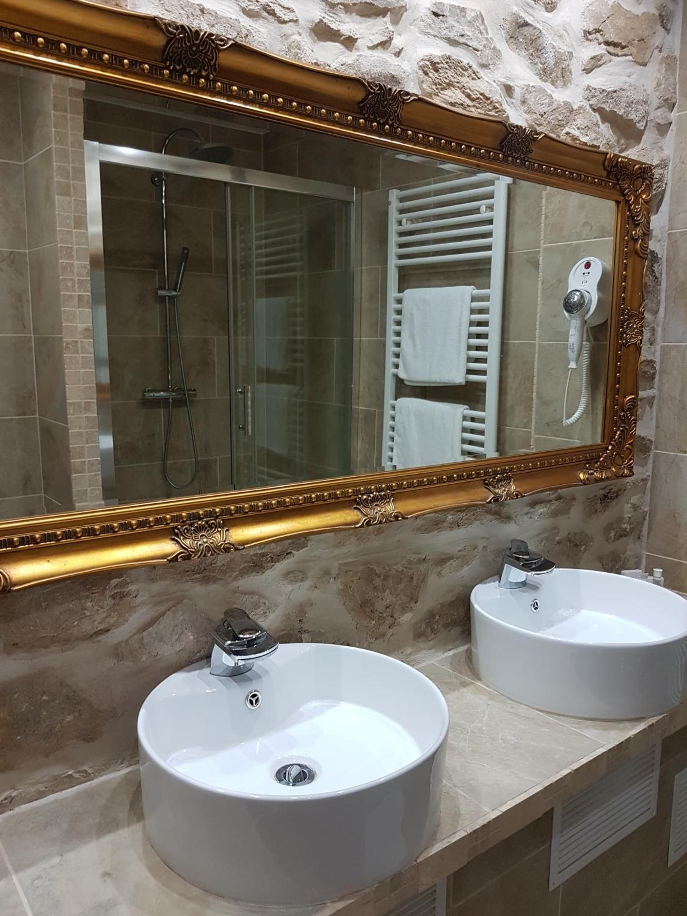 A bathroom with a large ornate mirror, two round sinks, a glass-enclosed shower, and a towel rack.