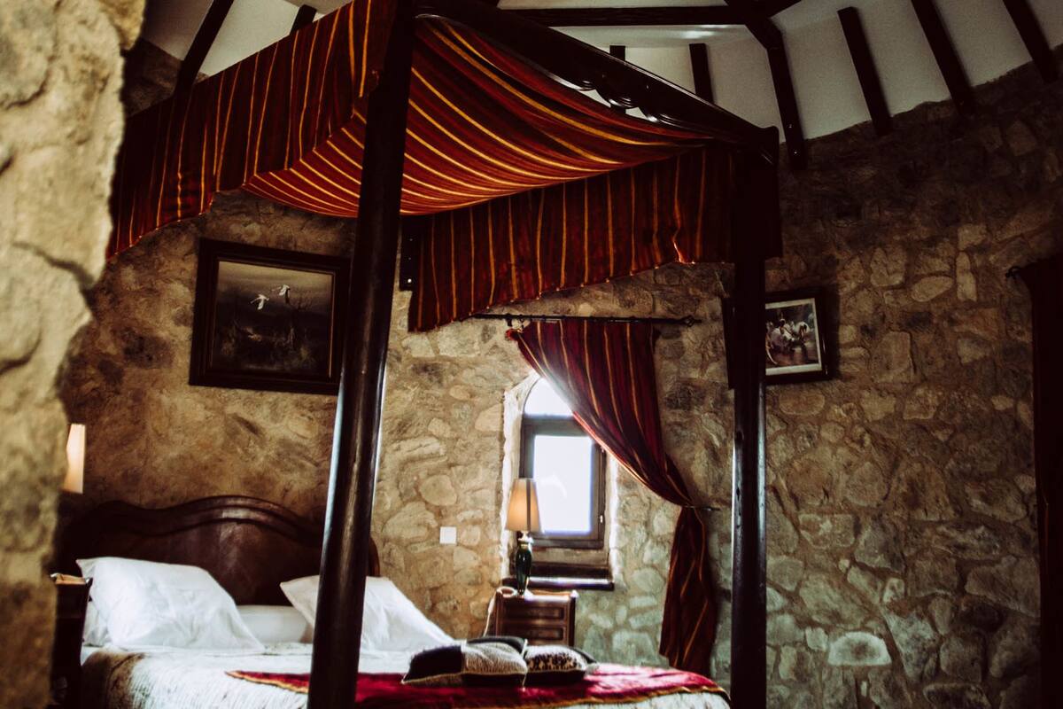 Une pièce avec un mur en pierre, un plafond en bois orné d’un motif géométrique, un lit à baldaquin et une fenêtre avec des rideaux.