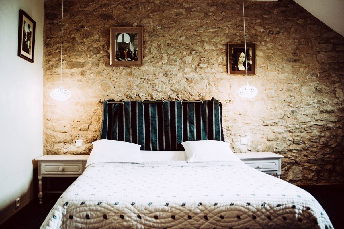 Une chambre avec un mur en pierre, un lit doté d’une tête de lit à rayures, deux tables de chevet et deux lampes suspendues.
