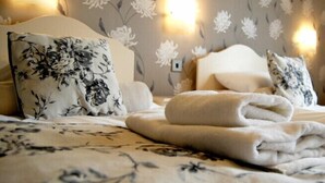 A neatly made bed with floral-patterned bedding and pillows, a folded white towel, and a wall with floral wallpaper and wall sconces. - Hillcrest Hotel (Lincoln)