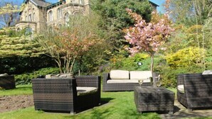 A garden with wicker furniture and a large building in the background. - Hillcrest Hotel (Lincoln)