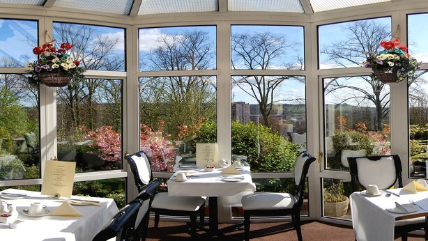 A conservatory with a table set for dining, large windows, and hanging flower baskets. - Hillcrest Hotel (Lincoln)