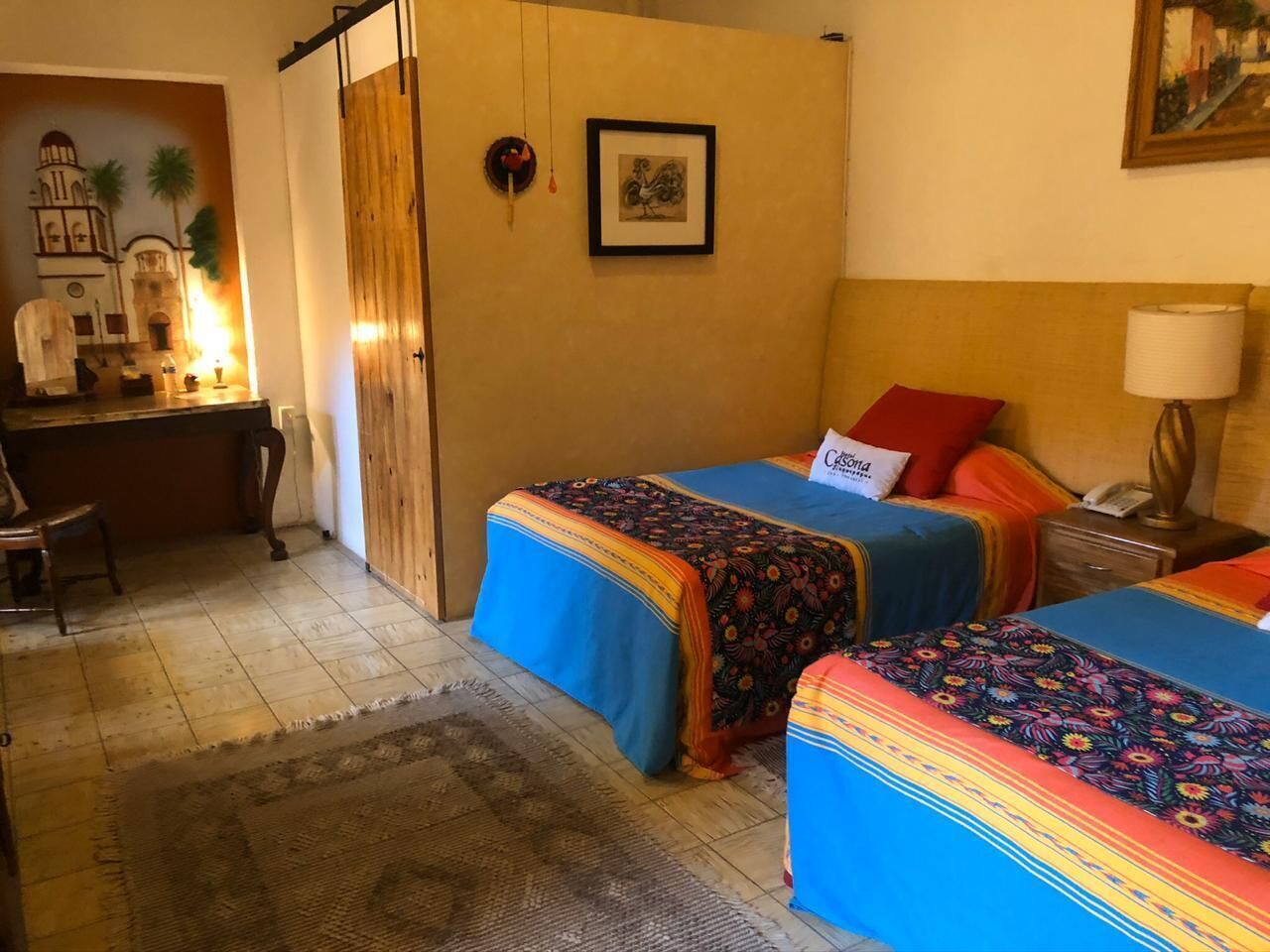A room with two beds, a desk, and a chair. There is a painting on the wall and a lamp on the desk.