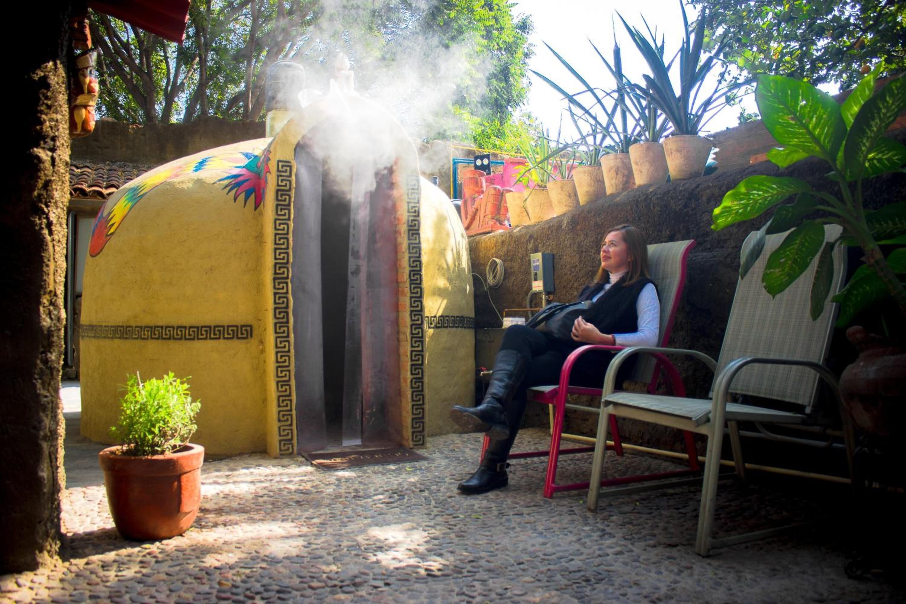 Une personne est assise sur une chaise dans une cour, près d’une structure décorative qui dégage de la vapeur, et est entourée de plantes en pot et d’un sol pavé.
