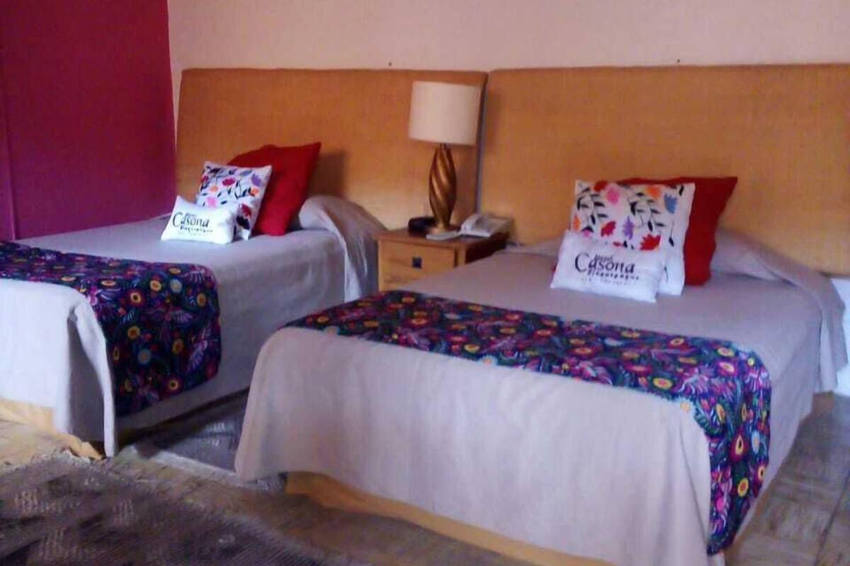 Une chambre d’hôtel avec deux lits simples, chacun recouvert d’une couvre-lit à motifs floraux et agrémenté d’un oreiller rouge. Une table de chevet avec une lampe et un téléphone se trouve entre les lits. Un tableau est accroché au mur au-dessus des lits, et une petite pancarte avec le mot « hôtel » est visible.