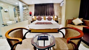 Quarto de hotel com uma cama grande, duas poltronas, uma mesa pequena e um painel de vidro.