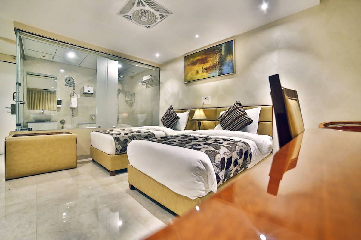 A modern hotel room with two beds, a glass shower enclosure, a bedside table, and a large painting on the wall.