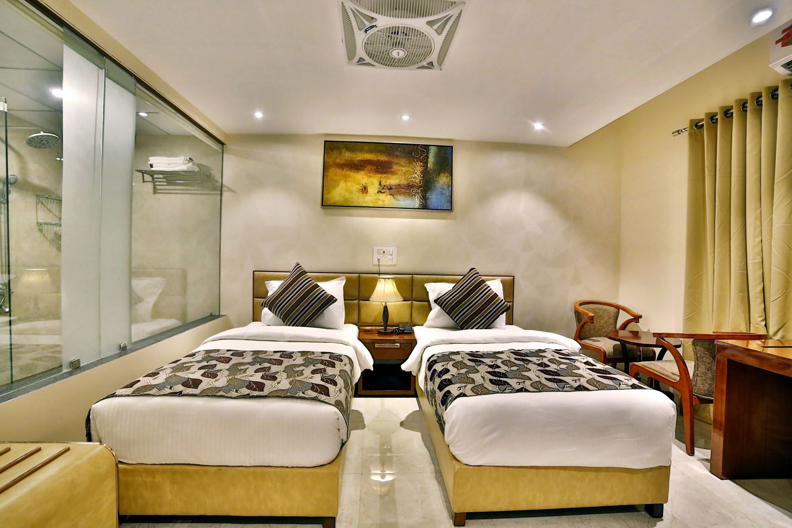 A hotel room with two beds, a desk, a chair, a glass shower enclosure, and a ceiling fan.