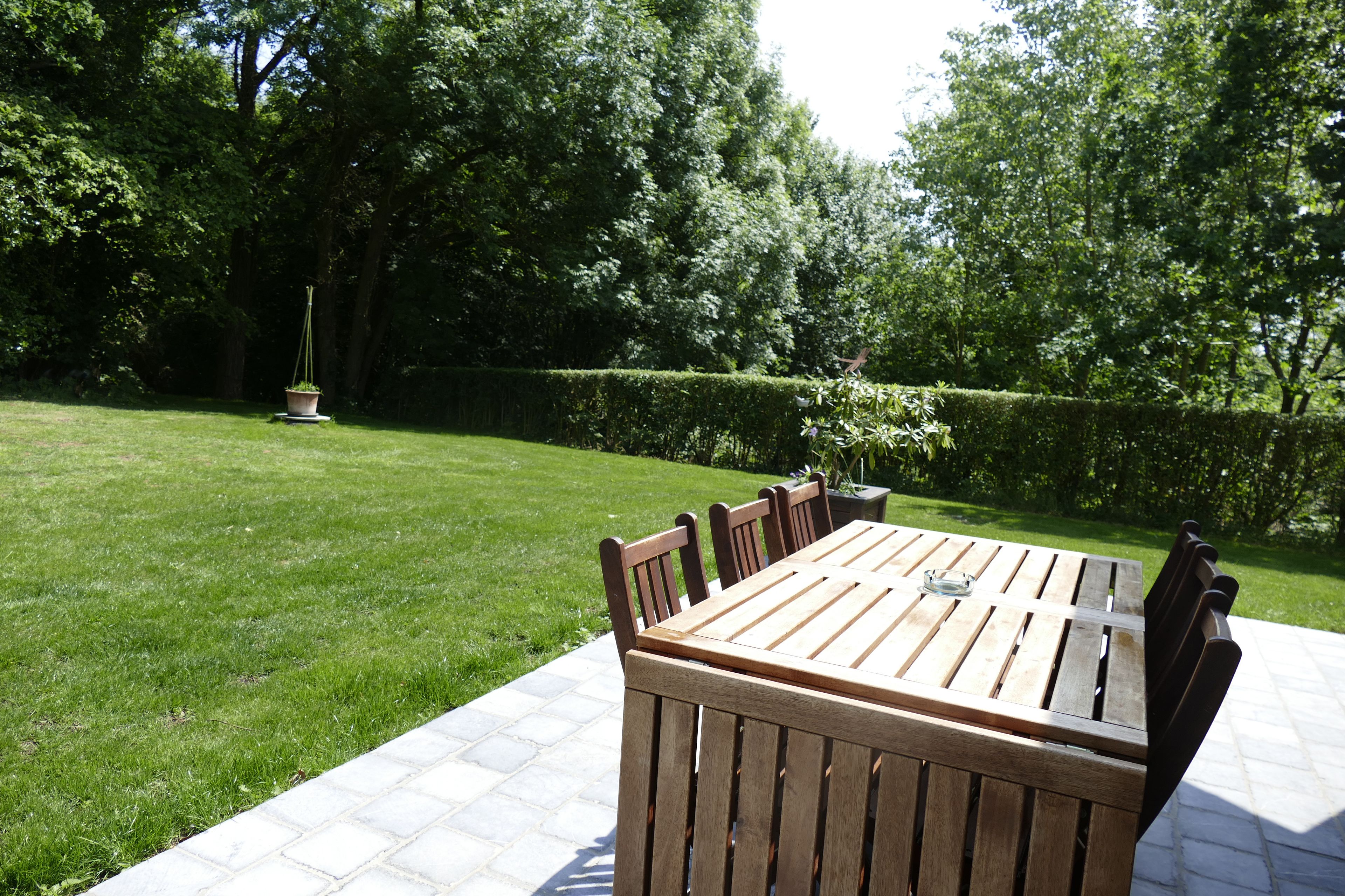 A wooden table and chairs set on a patio with a grassy lawn and hedges in the background.