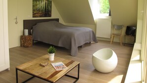 A bedroom with a bed, a bedside table with a plant, a chair, a TV, and a window. - La Garonde (Berthen)